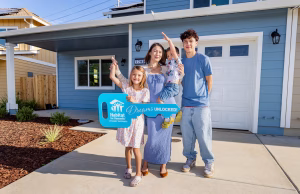 A family of four stands in front of their new Habitat home, smiling and holding a large blue Habitat key that says “Dreams Unlocked.”