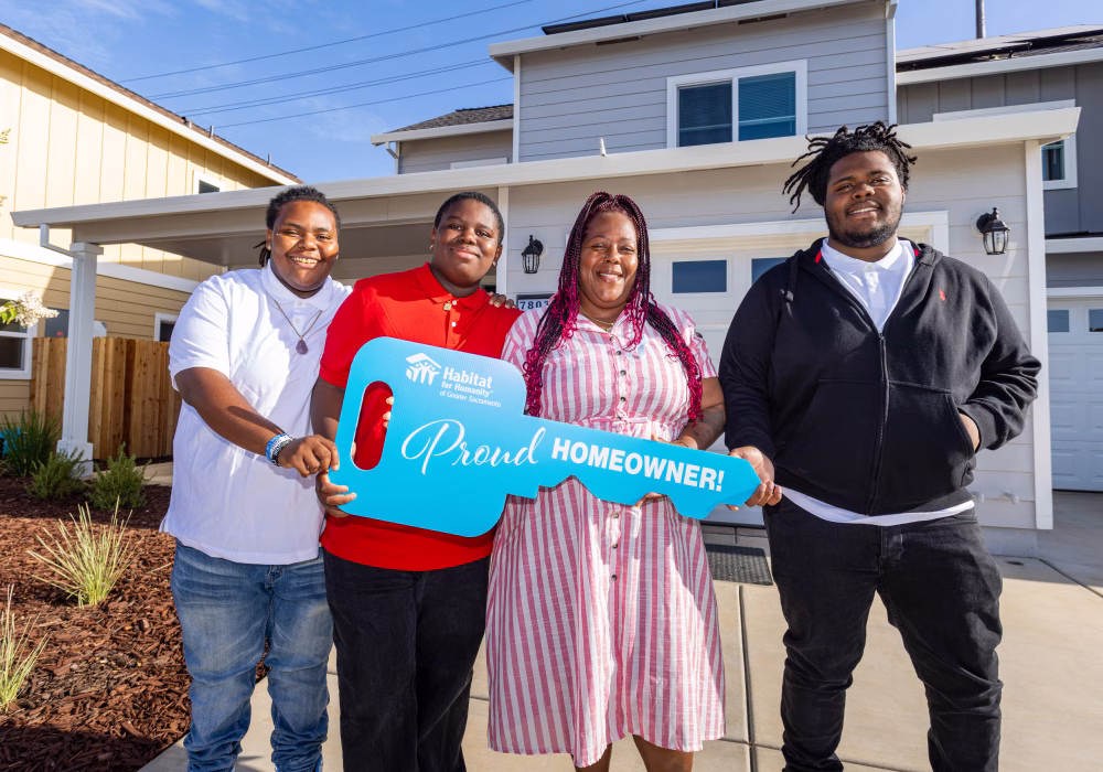 A smiling family of four stands in front of their new Habitat home while holding a large blue key that says “Proud Homeowner.”