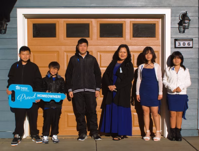 A family of six stands outside their new Habitat home in front of the garage, with one child holding a large blue key that says “Proud Homeowner.”