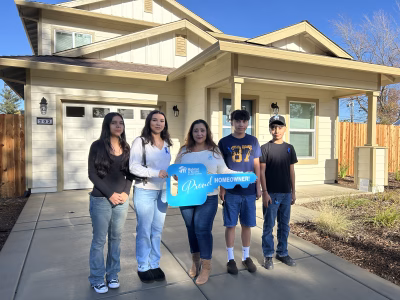 A family of five stands in front of their new Habitat home, with two family members holding a large blue Habitat key that says “Proud Homeowner.”