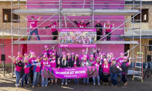 A large group of Habitat Women Build volunteers in pink shirts and hard hats gathers in front of a construction site with scaffolding and banners.
